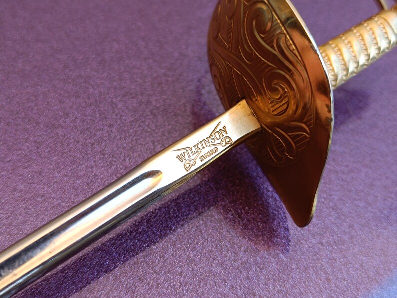 K&ouml;nnte beinhalten: Ein silbernes Schwert mit einem goldfarbenen Griff und einer Parierstange. Die Parierstange hat ein kompliziertes Design und die Klinge ist mit "WILKINSON SWORD" graviert.