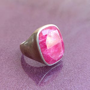 Op de afbeelding: Een zilveren ring met een grote, rechthoekige, gefacetteerde roze edelsteen. De ring heeft een eenvoudig, klassiek ontwerp met een gepolijste zilveren band. De edelsteen heeft een levendige roze kleur en vangt het licht.