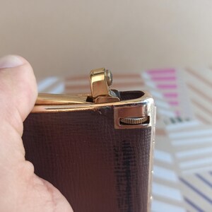 May include: A brown leather cigarette lighter with a gold-colored top and a small gold-colored knob on the side.