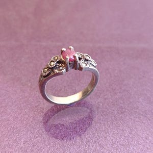 K&ouml;nnte beinhalten: Ein silberner Ring mit einem rosa Edelstein. Der Ring hat florale Details am Band und um den zentralen Stein. Der Ring ist vor einem lila Hintergrund platziert.