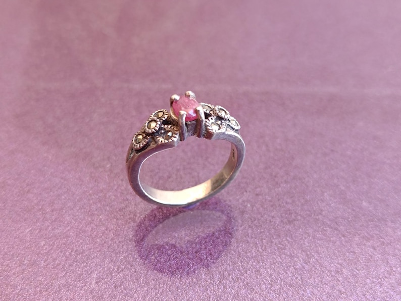 K&ouml;nnte beinhalten: Ein silberner Ring mit einem rosa Edelstein. Der Ring hat florale Details am Band und um den zentralen Stein. Der Ring ist vor einem lila Hintergrund platziert.