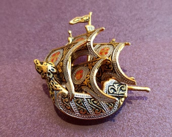 May include: A gold-toned ship brooch with intricate detailing. The brooch features a detailed ship design with red and gold accents, a small flag, and ornate patterns. The ship's sails and hull are embellished with decorative elements.