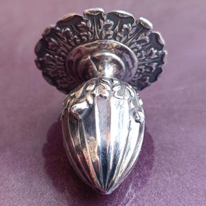 Może przedstawiać: Srebrny korek do butelki o bogatym wzornictwie. Korek ma szczegółową, żłobkowaną podstawę i dekoracyjną górę z kwiatowym wzorem. Obiekt umieszczony jest na fioletowym tle.