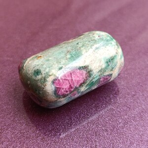 May include: A rounded, tumbled gemstone. The stone has a mottled green and white surface with striking red inclusions. The polished stone is set against a purple background, accentuating its natural colours and textures.