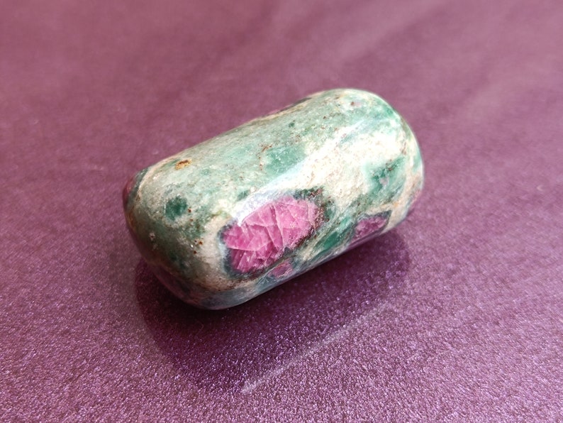 May include: A rounded, tumbled gemstone. The stone has a mottled green and white surface with striking red inclusions. The polished stone is set against a purple background, accentuating its natural colours and textures.