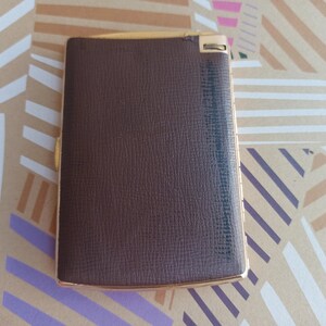 May include: A brown leather cigarette case with a gold trim and a sliding mechanism. The case has a textured surface and a gold metal hinge.
