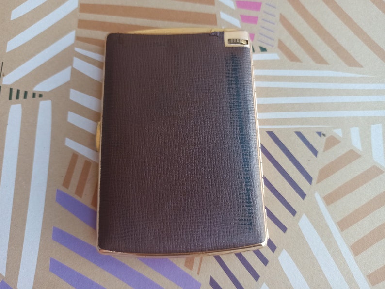 May include: A brown leather cigarette case with a gold trim and a sliding mechanism. The case has a textured surface and a gold metal hinge.