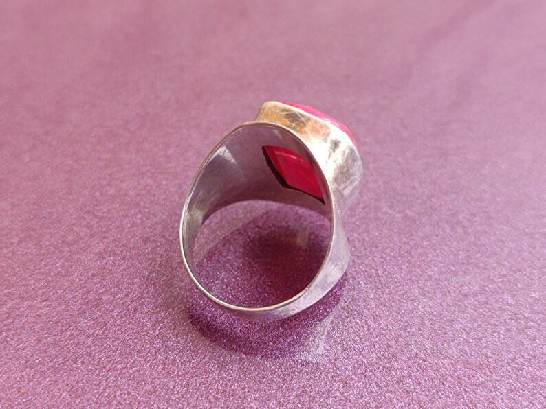 May include: A silver ring featuring a large, vivid red gemstone. The ring has a broad, curved band and a geometric shape to the gemstone. The gemstone is a bright, eye-catching colour, set in a silver bezel.