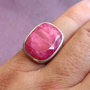 Op de afbeelding: Een grote, rechthoekige ring met een robijn edelsteen en een zilveren band. De robijn heeft een levendige roze kleur met zichtbare facetten. De ring wordt op een vinger gedragen.