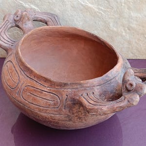 May include: A terracotta pot shaped like a turtle with two handles. The pot has a brown glaze and is decorated with incised lines.