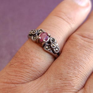 K&ouml;nnte beinhalten: Ein silberner Ring mit einem zentralen, runden, rosa Edelstein. Der Ring ist mit kleinen, dunklen, kreisf&ouml;rmigen Akzenten um den Edelstein und entlang des Bandes verziert. Der Ring befindet sich an einem Finger, vor einem violetten Hintergrund.