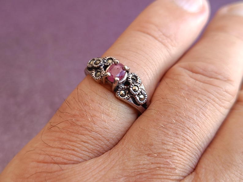 K&ouml;nnte beinhalten: Ein silberner Ring mit einem zentralen, runden, rosa Edelstein. Der Ring ist mit kleinen, dunklen, kreisf&ouml;rmigen Akzenten um den Edelstein und entlang des Bandes verziert. Der Ring befindet sich an einem Finger, vor einem violetten Hintergrund.