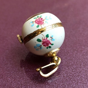 May include: A small, round, white trinket box with a gold clasp and accents. The box is decorated with pink roses and blue leaves. The box has a gold-colored knob and a gold-colored clasp.