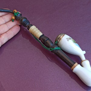 May include: A wooden pipe with a white porcelain bowl. The bowl has a painted scene of a landscape with trees and water. The pipe has a black mouthpiece and a green cord with beads.