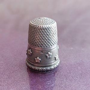 May include: A silver thimble with a textured surface and star-shaped embellishments. The thimble is designed for sewing and crafting, offering protection for the fingertip. The thimble is made of metal and has a classic, vintage appearance.