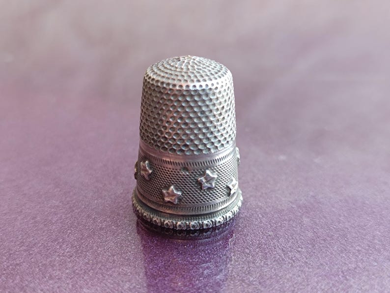 May include: A silver thimble with a textured surface and star-shaped embellishments. The thimble is designed for sewing and crafting, offering protection for the fingertip. The thimble is made of metal and has a classic, vintage appearance.