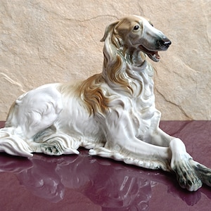 Op de afbeelding: Een porseleinen beeldje van een witte en bruine jachthond die ligt met zijn hoofd naar de zijkant gedraaid. De hond heeft een lang, slank lichaam en een lange, puntige snuit.