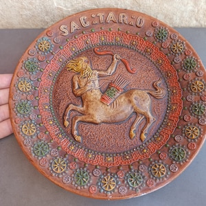 May include: A brown ceramic plate featuring a raised image of a centaur archer, surrounded by a colourful border of geometric patterns. The plate is inscribed with the word "SAGITTARIO".