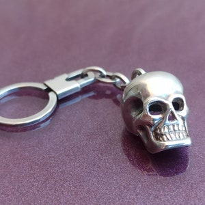 May include: A silver skull keychain with a detailed design. The skull has a chain attached to a silver ring.