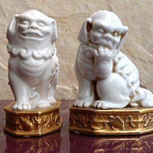 Op de afbeelding: Een paar witte keramische foo-honden, traditionele Chinese beschermleeuwen, met gouden accenten op hun bases. De foo-honden staan en zitten.