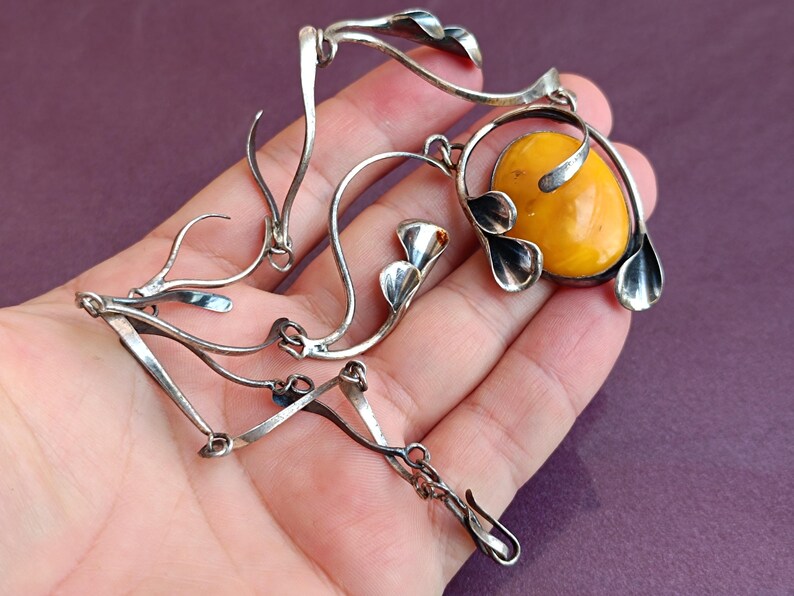May include: A silver necklace with a large amber stone pendant. The pendant is surrounded by silver leaves and the chain is made of silver links.