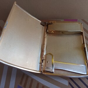 May include: A gold-colored metal cigarette case with a built-in lighter. The case is open, revealing a textured interior and a silver-colored lighter mechanism.