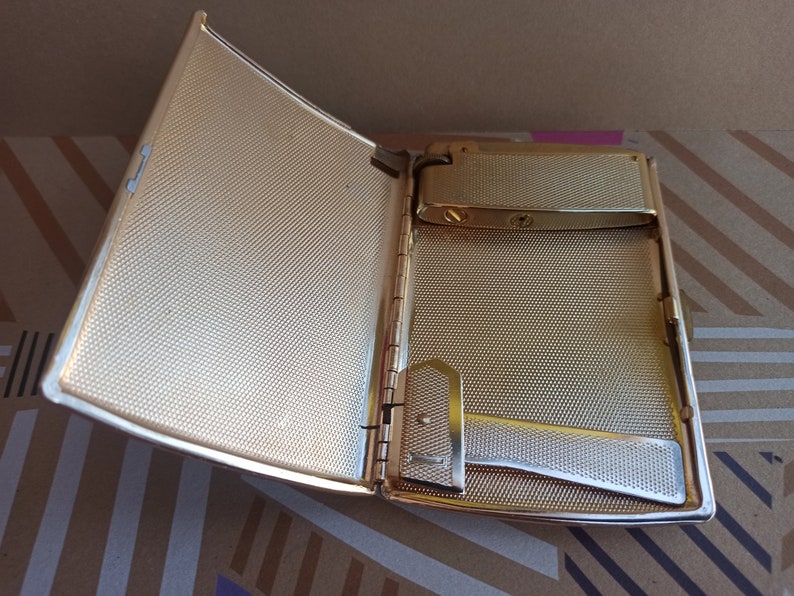 May include: A gold-colored metal cigarette case with a built-in lighter. The case is open, revealing a textured interior and a silver-colored lighter mechanism.