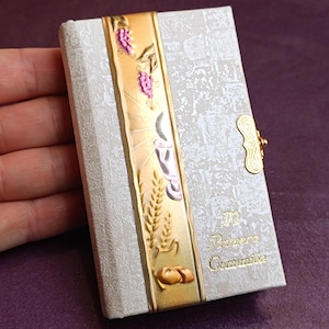 May include: A small, rectangular, off-white book with a gold clasp. The cover features embossed patterns and a gold band with religious symbols and the words "Primera Comunión". The book is held in a hand.