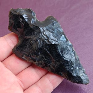 Op de afbeelding: Een zwarte obsidianen pijlpunt met een puntige punt en een licht gebogen basis. De pijlpunt is glad en gepolijst, met een glanzende afwerking.