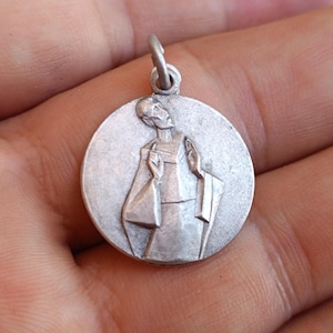 May include: A silver-coloured round pendant featuring a detailed relief of a robed figure with raised arms. The pendant has a small loop at the top for a chain. The design is vintage and detailed.