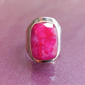 May include: A silver ring with a large, rectangular, faceted ruby gemstone. The ruby is a vibrant pink-red. The ring has a simple, modern design with a wide band.