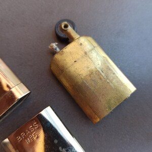 May include: A vintage brass lighter with a black wheel and a brass case. The lighter has a small wick and a brass cap. The lighter is marked "BRASS NPS".