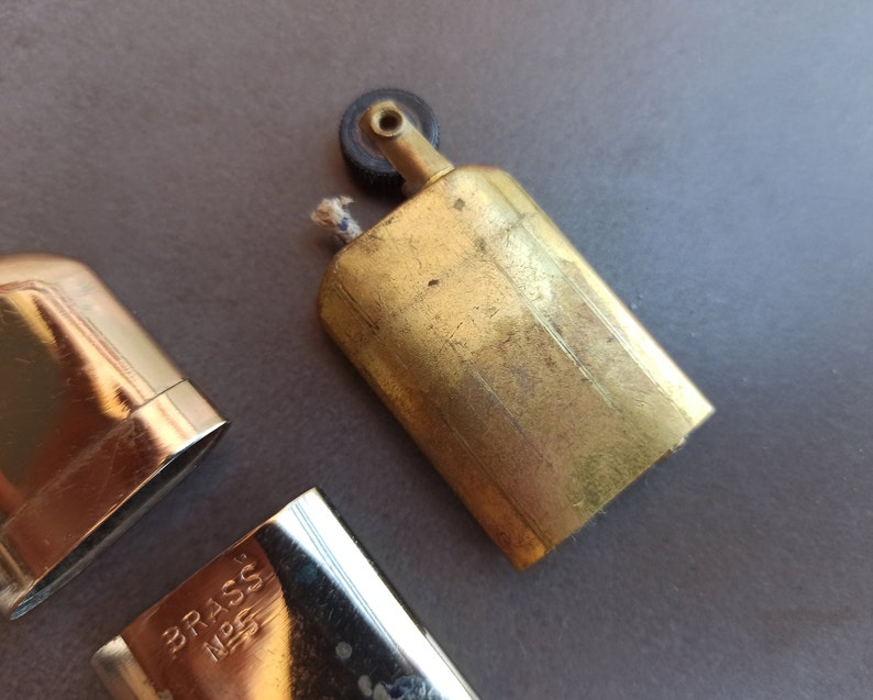 May include: A vintage brass lighter with a black wheel and a brass case. The lighter has a small wick and a brass cap. The lighter is marked "BRASS NPS".