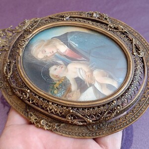 K&ouml;nnte beinhalten: Ein antiker ovaler religi&ouml;ser Bilderrahmen mit detaillierter Bronzeoberfl&auml;che. Der Rahmen umgibt ein Gem&auml;lde einer Frau und eines Kindes. Der Rahmen hat ein verziertes, filigranes Design um das ovale Gem&auml;lde.