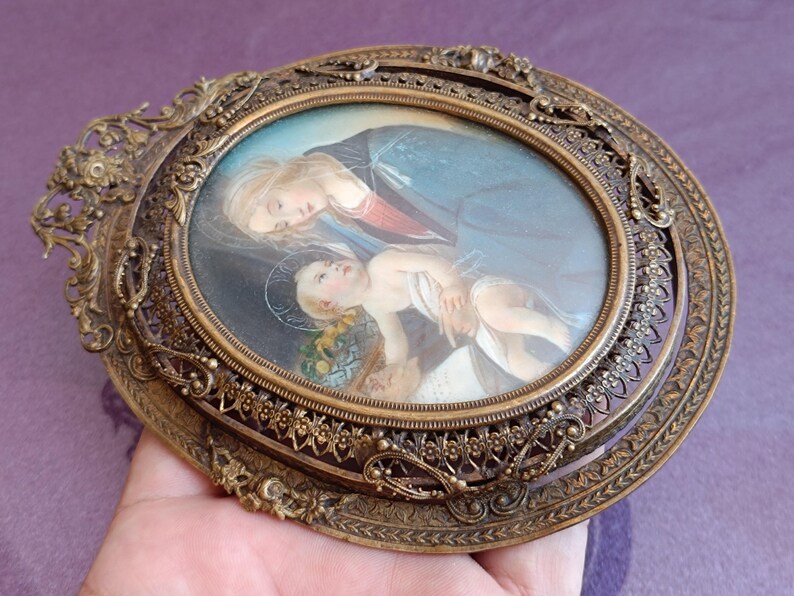 K&ouml;nnte beinhalten: Ein antiker ovaler religi&ouml;ser Bilderrahmen mit detaillierter Bronzeoberfl&auml;che. Der Rahmen umgibt ein Gem&auml;lde einer Frau und eines Kindes. Der Rahmen hat ein verziertes, filigranes Design um das ovale Gem&auml;lde.
