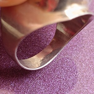 May include: Close-up of a silver ring with a smooth, polished surface. The ring has a wide band and a slightly curved design. The inside of the ring is visible, with markings that appear to be a hallmark. The ring is on a purple surface.