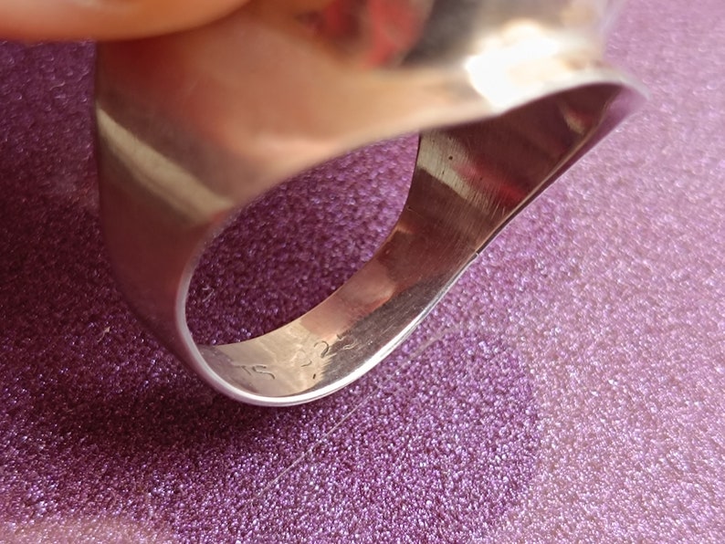 May include: Close-up of a silver ring with a smooth, polished surface. The ring has a wide band and a slightly curved design. The inside of the ring is visible, with markings that appear to be a hallmark. The ring is on a purple surface.