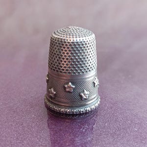 May include: A silver thimble with a textured surface and a band of embossed stars. The thimble is a sewing tool, designed to protect the finger while pushing a needle through fabric. The thimble is approximately 1 inch tall.