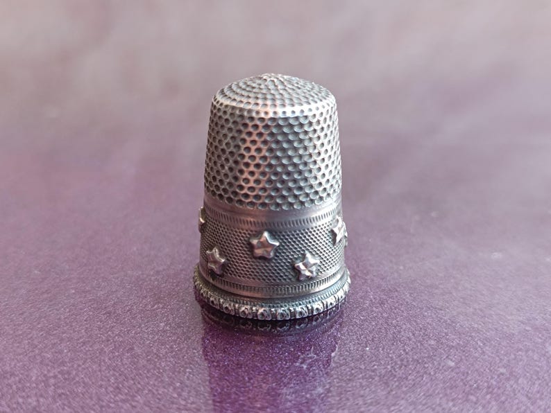 May include: A silver thimble with a textured surface and a band of embossed stars. The thimble is a sewing tool, designed to protect the finger while pushing a needle through fabric. The thimble is approximately 1 inch tall.