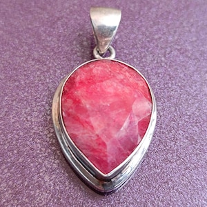 Può includere: Un ciondolo a forma di goccia con una grande pietra preziosa rosa sfaccettata incastonata in una cornice di metallo color argento. Il ciondolo ha un anello nella parte superiore per una catena. Lo sfondo è viola strutturato.