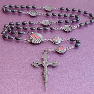May include: A vintage rosary with dark beads and silver-toned metal accents. The rosary features decorative pieces with red cross designs and a detailed crucifix pendant. The beads are oval-shaped and strung together.