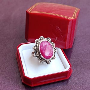Op de afbeelding: Een zilveren ring met een grote, ovale, roze edelsteen, gezet in een decoratieve bezel. De ring wordt gepresenteerd in een open rode sieradendoos met een wit interieur en gouden rand.
