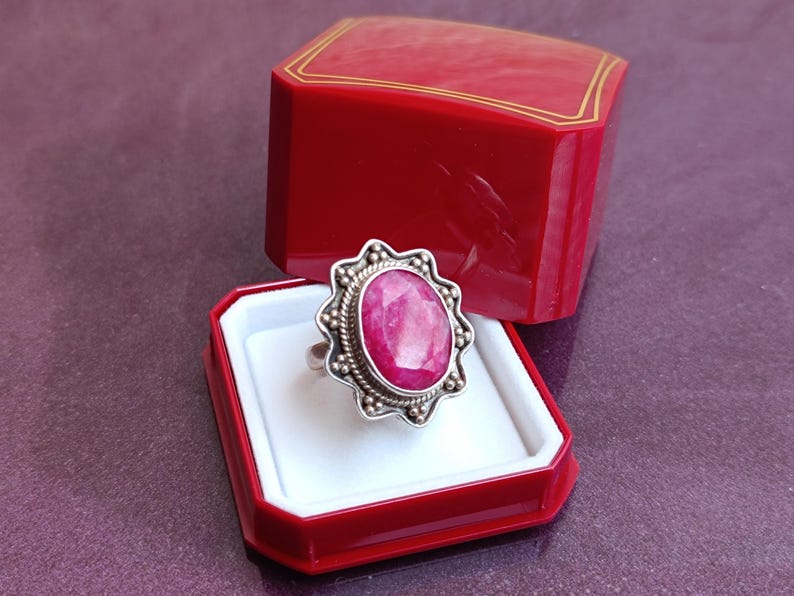 Op de afbeelding: Een zilveren ring met een grote, ovale, roze edelsteen, gezet in een decoratieve bezel. De ring wordt gepresenteerd in een open rode sieradendoos met een wit interieur en gouden rand.