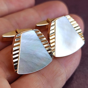 May include: A pair of gold-toned cufflinks featuring a trapezoidal shape with a central inlay of iridescent white mother-of-pearl. The sides of each cufflink are accented with a series of parallel, raised gold-toned ridges along the edges.