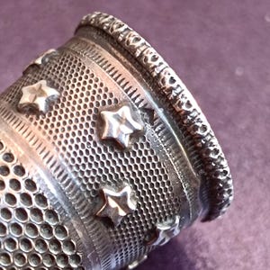 May include: Close-up of a silver thimble with a detailed design. The thimble features a series of star-shaped embellishments and a textured surface. The top edge has a decorative pattern. The thimble is a sewing tool.