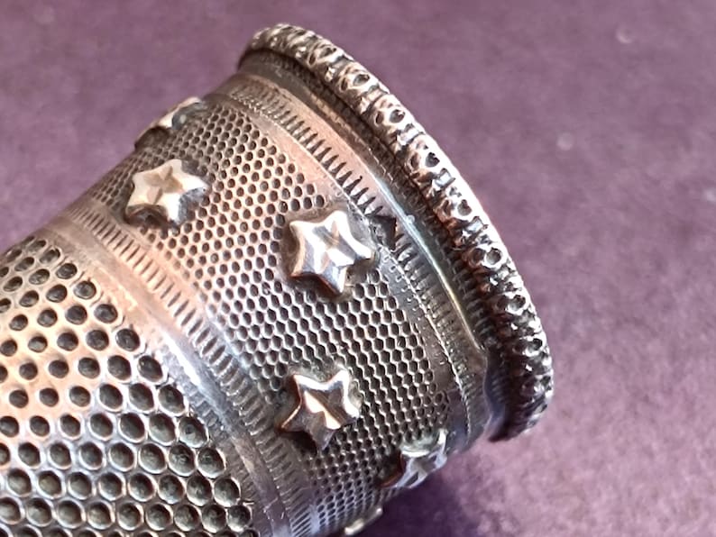 May include: Close-up of a silver thimble with a detailed design. The thimble features a series of star-shaped embellishments and a textured surface. The top edge has a decorative pattern. The thimble is a sewing tool.