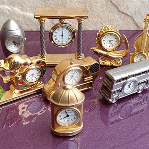 May include: A collection of miniature decorative clocks in various shapes and styles. The clocks include a rocket, a double-decker bus, a stand-alone clock, and clocks shaped like an elephant, a musical instrument, and a decorative stand. The clocks are gold and silver.