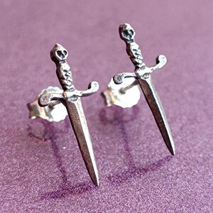 May include: A pair of silver-toned dagger stud earrings. Each earring features a detailed dagger design with a skull on the hilt. The earrings are set against a purple background, showcasing their intricate craftsmanship.
