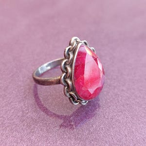 Pode incluir: Um anel de prata com uma gema vermelha em forma de l&aacute;grima engastada em uma moldura de prata decorativa.