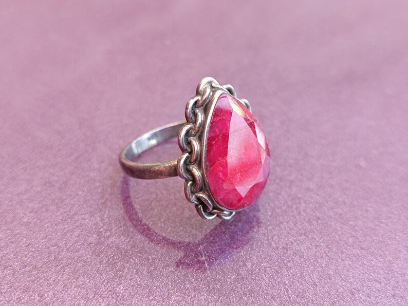 Pode incluir: Um anel de prata com uma gema vermelha em forma de l&aacute;grima engastada em uma moldura de prata decorativa.